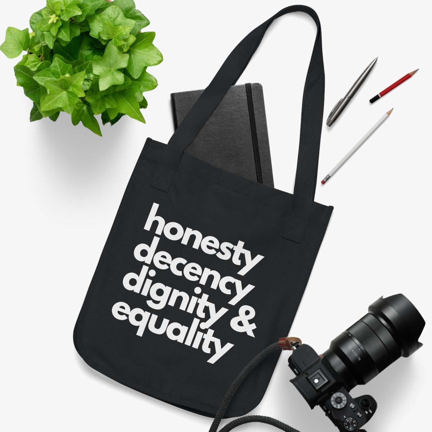 Black tote bag with 'honesty decency dignity & equality' text on a white surface with a plant, book, and camera.