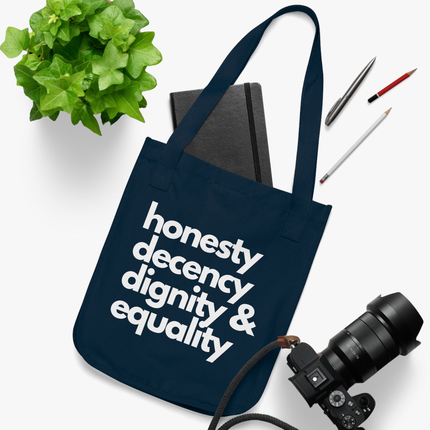 Navy tote bag with 'honesty decency dignity & equality' text on a white surface with a plant, notebook, and camera.